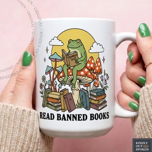 May include: White ceramic mug featuring a green frog reading a book titled "Banned Tales" while sitting on a mushroom. The mug has a whimsical design with mushrooms, books, and the text "READ BANNED BOOKS".