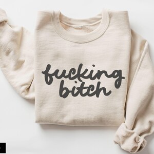 Puede incluir: Sudadera color arena con la frase "fucking bitch" en escritura gris oscuro. Las mangas est&aacute;n enrolladas. La palabra "SAND" est&aacute; en un rect&aacute;ngulo negro en la esquina inferior izquierda. La sudadera est&aacute; doblada.