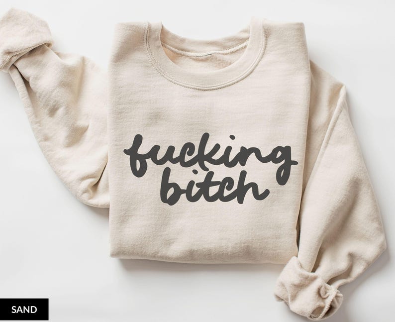 Puede incluir: Sudadera color arena con la frase "fucking bitch" en escritura gris oscuro. Las mangas est&aacute;n enrolladas. La palabra "SAND" est&aacute; en un rect&aacute;ngulo negro en la esquina inferior izquierda. La sudadera est&aacute; doblada.