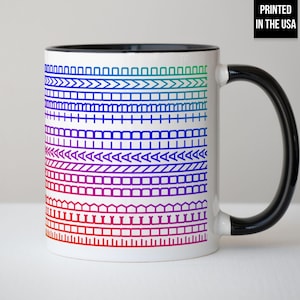 Op de afbeelding: Witte keramische mok met een zwarte handgreep en binnenkant. De mok heeft een kleurrijk, geometrisch patroon in rood, paars, blauw en groen. De tekst "PRINTED IN THE USA" is zwart op een witte achtergrond gedrukt.