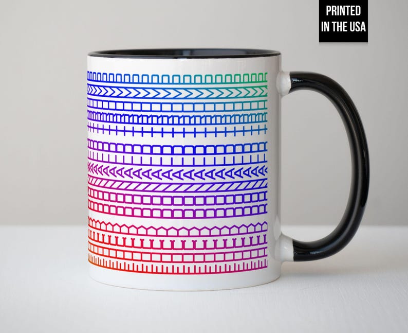 Op de afbeelding: Witte keramische mok met een zwarte handgreep en binnenkant. De mok heeft een kleurrijk, geometrisch patroon in rood, paars, blauw en groen. De tekst "PRINTED IN THE USA" is zwart op een witte achtergrond gedrukt.