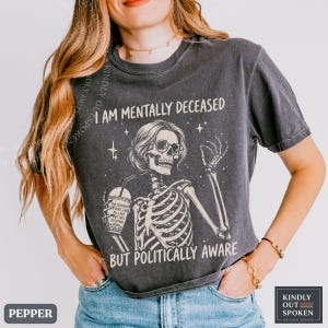 May include: A charcoal gray t-shirt featuring a graphic of a skeleton wearing glasses, holding a drink, and surrounded by stars. The text reads I AM MENTALLY DECEASED BUT POLITICALLY AWARE, with smaller text on the drink cup.