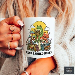 May include: White ceramic mug featuring a whimsical illustration of a frog reading a book titled "Banned Tales" while sitting on a mushroom. The mug also has the text "Read Banned Books" in black. The design includes a yellow sun, clouds, and other mushrooms.