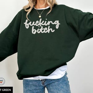 Puede incluir: Sudadera verde bosque con la frase "fucking bitch" en escritura blanca. La sudadera tiene mangas largas y cuello redondo. La modelo lleva una camiseta blanca y vaqueros azules.