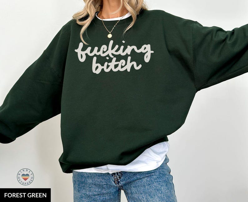 Puede incluir: Sudadera verde bosque con la frase "fucking bitch" en escritura blanca. La sudadera tiene mangas largas y cuello redondo. La modelo lleva una camiseta blanca y vaqueros azules.