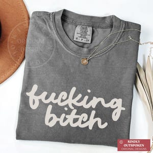 May include: A gray Comfort Colors t-shirt with the words "fucking bitch" in a cursive font. A silver necklace with a small pendant is draped across the shirt. The shirt is folded on a white surface.