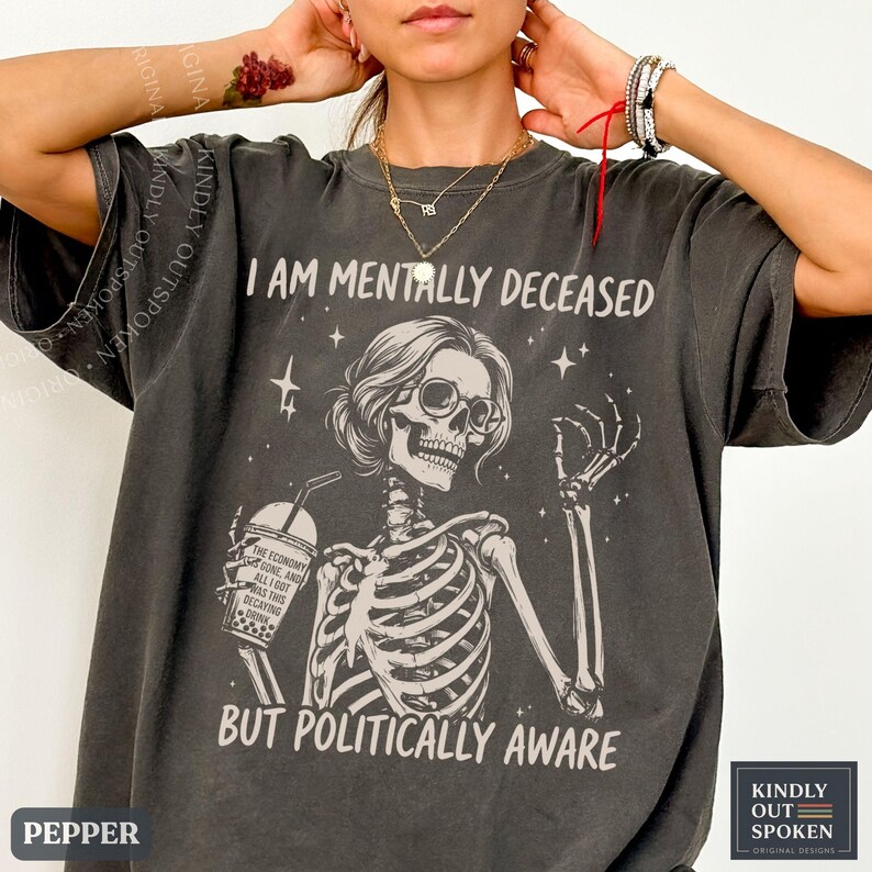 May include: A charcoal gray t-shirt featuring a graphic of a skeleton wearing glasses, holding a boba drink, and making an okay hand gesture. The text reads I AM MENTALLY DECEASED BUT POLITICALLY AWARE, with the drink cup labeled THE ECONOMY IS GONE.