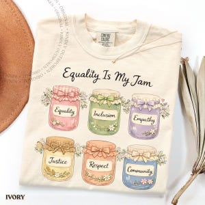 May include: An ivory t-shirt featuring the text Equality Is My Jam and six illustrated jars with bows and flowers. The jars are labeled Equality, Inclusion, Empathy, Justice, Respect, and Community. The shirt has a Comfort Colors brand tag.