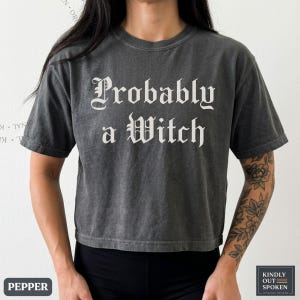 May include: A charcoal grey short-sleeve t-shirt with a crew neckline. The shirt features the white text "Probably a Witch" printed across the chest in a gothic-style font. The fabric has a washed, vintage-inspired texture and a cropped hemline.