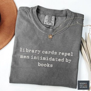 Może przedstawiać: Ciemnoszary t-shirt z krótkim rękawem i okrągłym dekoltem, z białym napisem "library cards repel men intimidated by books" nadrukowanym z przodu. Koszulka posiada metkę marki Comfort Colors przy wewnętrznej stronie kołnierza.