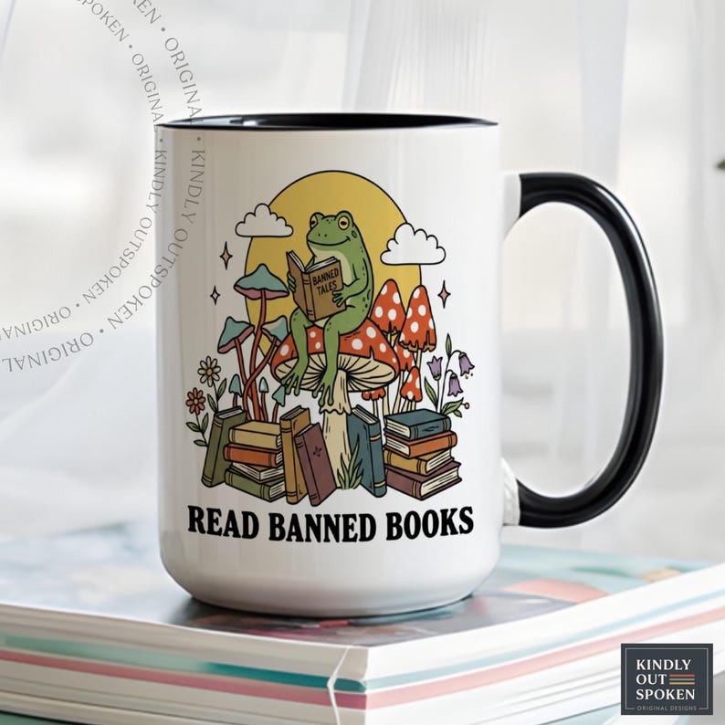 May include: White ceramic mug with a black handle and rim. The mug features a whimsical illustration of a frog reading a book titled "Banned Tales" while sitting on a mushroom. The text "READ BANNED BOOKS" is printed below the illustration.