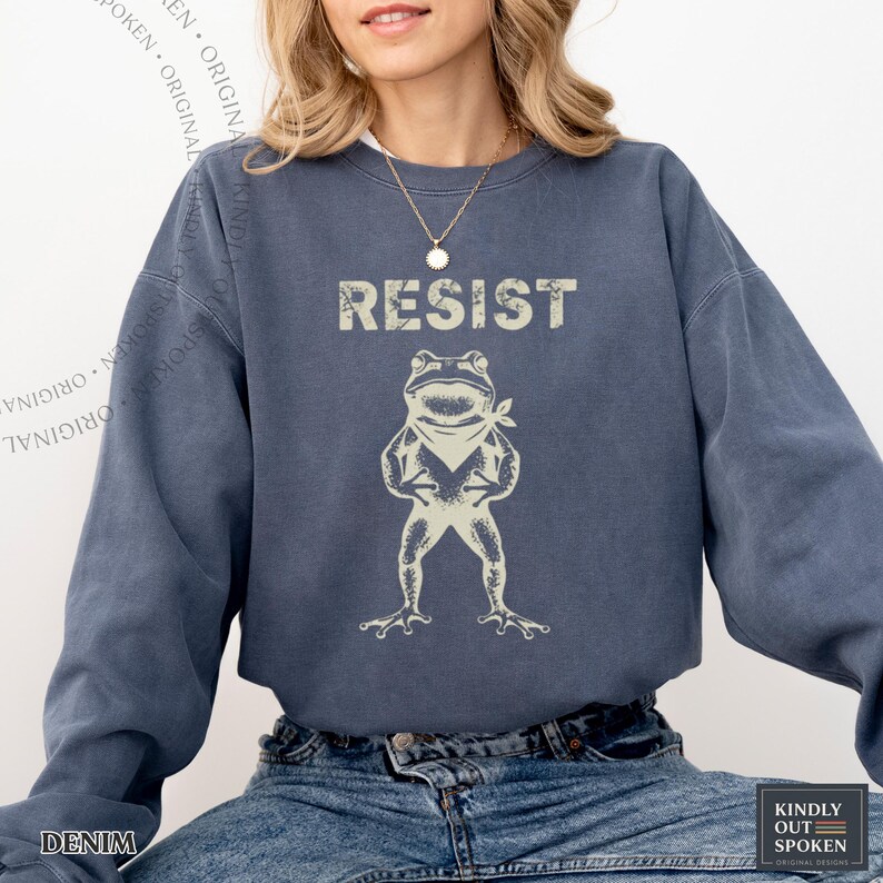 Op de afbeelding: Denimblauwe sweatshirt met het woord "RESIST" boven een cartoonkikker met een bandana. De sweatshirt heeft een relaxte pasvorm en lange mouwen. De tekst en de kikkergrafiek zijn in een vervaagde, vintage stijl.