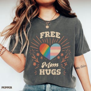 以下が含まれることがあります： チャコールグレーの半袖Tシャツ。中央にはレインボーカラーとトランスジェンダーフラッグのカラーで分割されたハートのグラフィックが描かれています。クリーム色の文字で「FREE Mom HUGS」と記され、周囲には太陽の光線と花のアクセントが添えられています。
