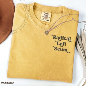 Puede incluir: Una camiseta de manga corta color amarillo mostaza con cuello redondo y etiqueta de la marca Comfort Colors. La prenda tiene el texto negro Radical Left Scum impreso en el lado derecho. Un collar de cadena plateada con un dije de rosa descansa encima.