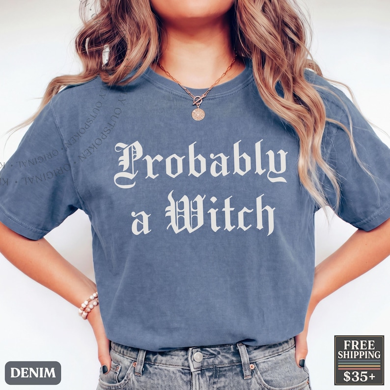 May include: A blue short-sleeve t-shirt featuring the white text "Probably a Witch" in a gothic-style font printed across the chest. The garment has a crew neckline and a relaxed fit, constructed from a solid-colored fabric with a subtle texture.