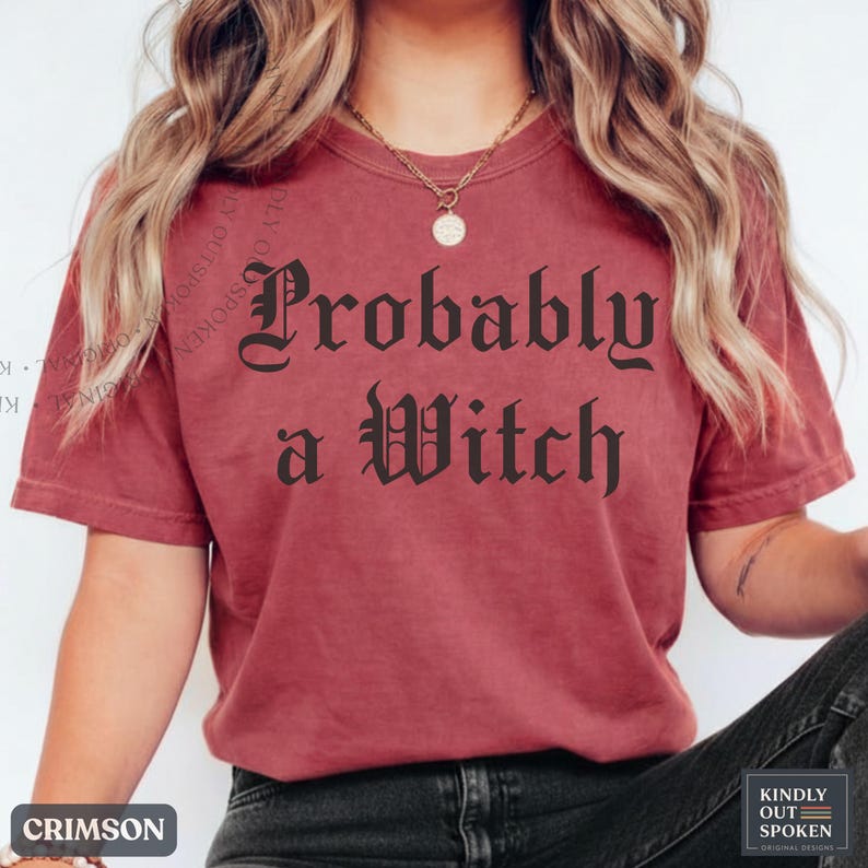 May include: A crimson short-sleeved t-shirt featuring the black text "Probably a Witch" in a gothic-style font centered on the chest. The garment has a crew neckline and a relaxed fit, paired with a gold-toned chain necklace with a round pendant.