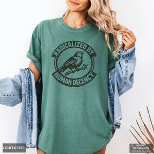 May include: A light green short-sleeved t-shirt featuring a black circular graphic of a bird perched on a branch. The text "RADICALIZED BY HUMAN DECENCY" is printed in a circular pattern around the bird. The shirt is worn with a blue denim jacket.