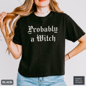 May include: A black short-sleeved t-shirt featuring the white text 'Probably a Witch' printed in a gothic-style font across the chest. The garment has a crew neckline and a standard fit, paired with blue denim jeans.