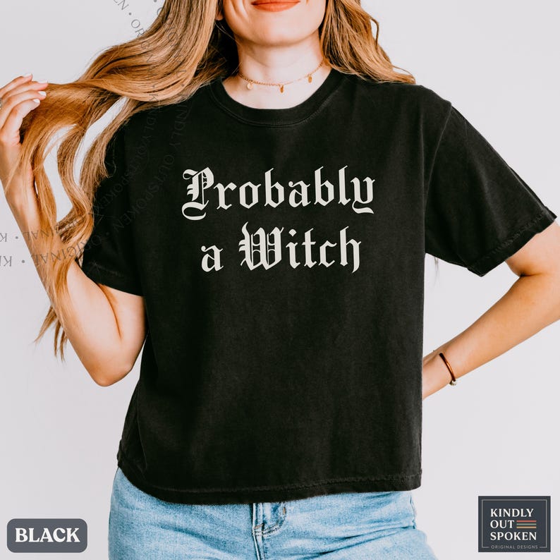 May include: A black short-sleeved t-shirt featuring the white text 'Probably a Witch' printed in a gothic-style font across the chest. The garment has a crew neckline and a standard fit, paired with blue denim jeans.