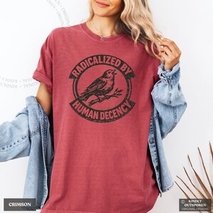 May include: A crimson short-sleeve t-shirt featuring a black circular graphic with a bird perched on a branch in the center. The text RADICALIZED BY HUMAN DECENCY is printed in a bold, distressed font around the circular border of the design.