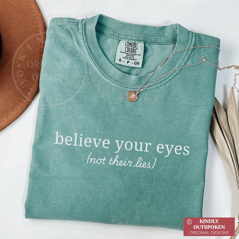 Pu&ograve; includere: Una t-shirt a maniche corte color verde salvia con scollo rotondo e la scritta bianca "believe your eyes (not their lies)". La maglia presenta un'etichetta del marchio Comfort Colors all'interno del collo ed &egrave; abbinata a una catenina dorata.