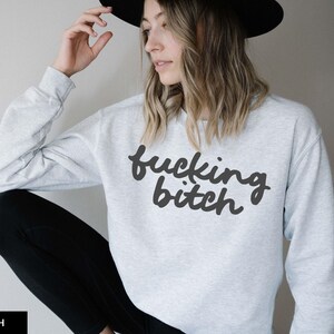 Puede incluir: Sudadera gris ceniza con el texto "fucking bitch" en negro, en fuente cursiva. La sudadera de manga larga se combina con pantalones negros. Se lleva un sombrero negro.