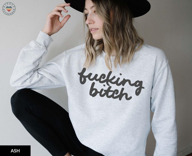 Puede incluir: Sudadera gris ceniza con el texto "fucking bitch" en negro, en fuente cursiva. La sudadera de manga larga se combina con pantalones negros. Se lleva un sombrero negro.