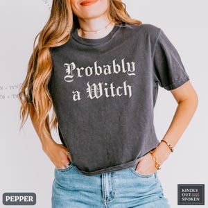 May include: A charcoal gray short-sleeve t-shirt featuring the white text "Probably a Witch" in a gothic-style font. The shirt has a crew neckline and a relaxed fit, paired with blue denim jeans. The design is centered on the chest area.