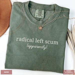 May include: A moss green short-sleeve t-shirt featuring the white text "radical left scum (apparently)" printed on the chest. The shirt has a crew neckline and a Comfort Colors brand label inside the collar, indicating a size small.