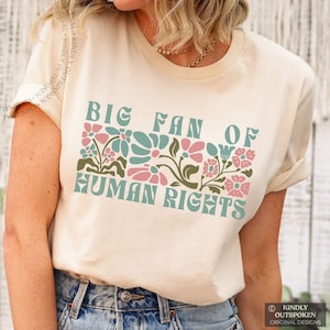 May include: Cream-colored t-shirt with the text "BIG FAN OF HUMAN RIGHTS" in a retro font. The text is above a floral design in pink, teal, and green. The shirt is a casual style.