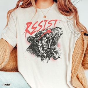 May include: An ivory short-sleeve t-shirt featuring a black graphic illustration of a roaring panther head. Above the panther, the word RESIST is printed in a distressed red font. A small red star is positioned near the panther's eye.