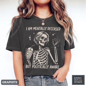 May include: A graphite-colored t-shirt featuring a graphic of a skeleton wearing sunglasses and holding a drink. The text reads I AM MENTALLY DECEASED BUT POLITICALLY AWARE. The drink cup has text that reads THE ECONOMY IS GONE AND ALL I GOT WAS THIS DECAYING DRINK.