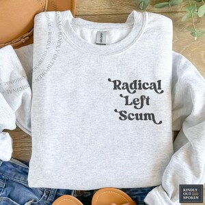 Puede incluir: Sudadera gris claro con la frase "Radical Left Scum" en negro con una fuente estilizada. La sudadera es de cuello redondo y parece estar hecha de un material suave. La etiqueta de la marca "Gildan" es visible.