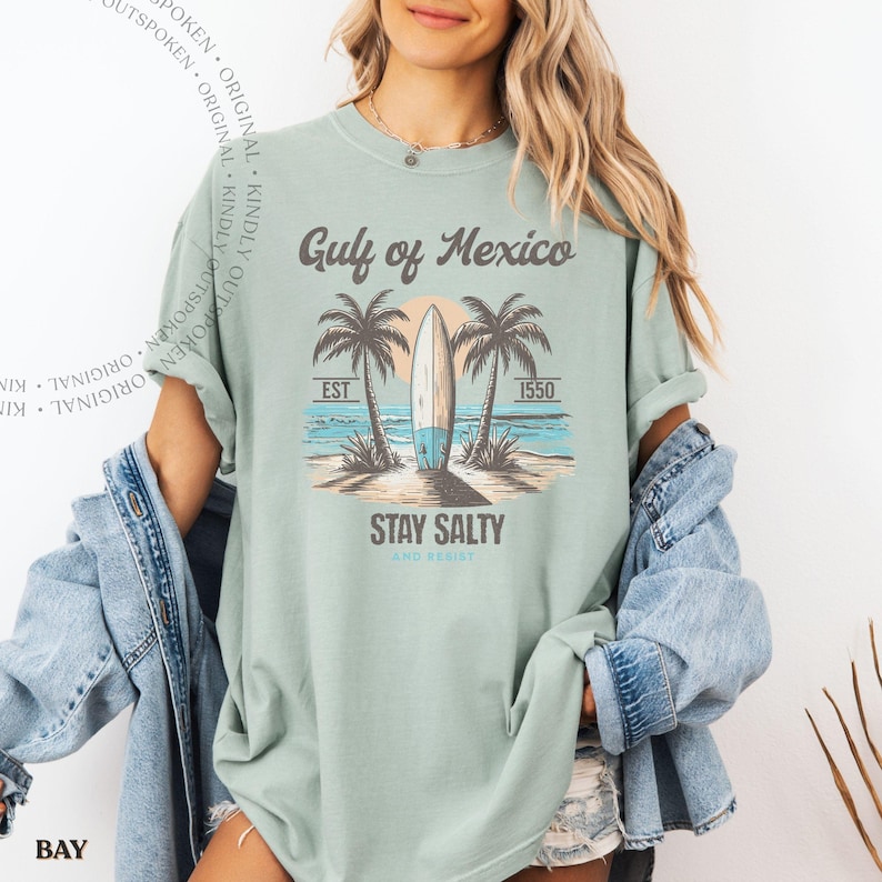May include: A sage green short-sleeve t-shirt featuring a graphic print of a surfboard on a beach with palm trees, the text Gulf of Mexico, EST 1550, and STAY SALTY AND RESIST. The shirt is worn with a blue denim jacket over the shoulders.