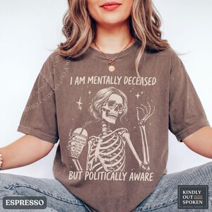 May include: A brown short-sleeve t-shirt featuring a graphic of a skeleton wearing sunglasses, holding a drink, and surrounded by stars. The text reads I AM MENTALLY DECEASED BUT POLITICALLY AWARE, with additional text on the drink cup.