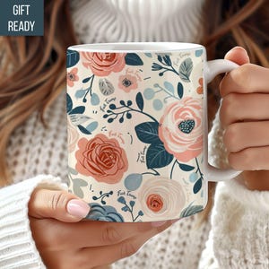May include: White ceramic mug featuring a floral design in pink, peach, and blue hues. The pattern includes roses and other flowers, with the text "Fuck Trump" interspersed. The mug is held by a person wearing a white sweater.