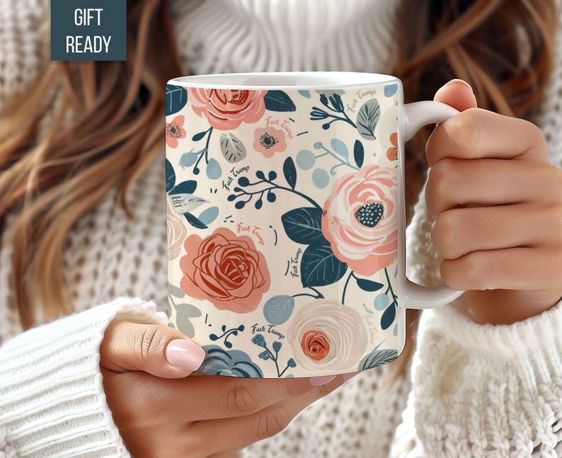 May include: White ceramic mug featuring a floral design in pink, peach, and blue hues. The pattern includes roses and other flowers, with the text "Fuck Trump" interspersed. The mug is held by a person wearing a white sweater.