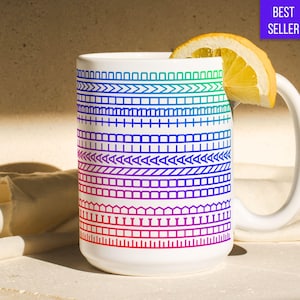 Op de afbeelding: Witte keramische mok met een kleurrijk geometrisch patroon in tinten blauw, groen en rood. Een schijfje citroen rust op de rand. De mok heeft een wit handvat en de woorden "BEST SELLER" staan in een paarse doos.