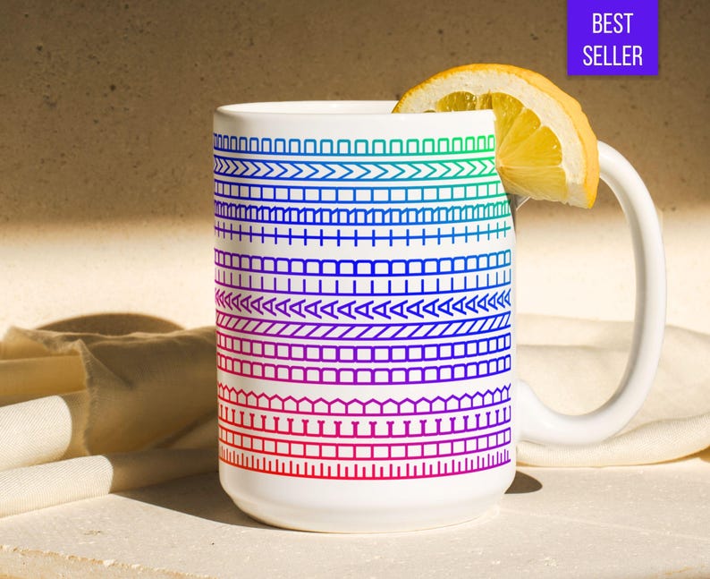 Op de afbeelding: Witte keramische mok met een kleurrijk geometrisch patroon in tinten blauw, groen en rood. Een schijfje citroen rust op de rand. De mok heeft een wit handvat en de woorden "BEST SELLER" staan in een paarse doos.