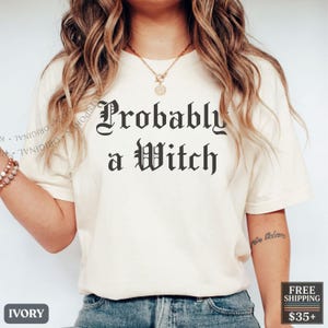 May include: An ivory short-sleeved t-shirt featuring the black text "Probably a Witch" in a gothic-style font centered on the chest. The shirt is paired with blue denim jeans and a gold-toned necklace with a circular pendant.
