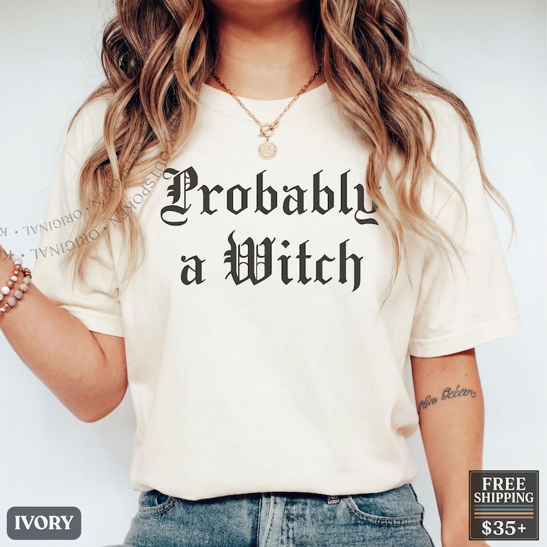 May include: An ivory short-sleeved t-shirt featuring the black text "Probably a Witch" in a gothic-style font centered on the chest. The shirt is paired with blue denim jeans and a gold-toned necklace with a circular pendant.