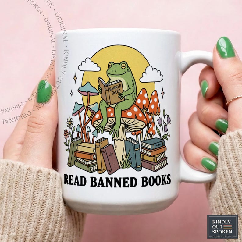 May include: White ceramic mug featuring a green frog reading a book titled "Banned Tales" while sitting on a mushroom. The mug has a whimsical design with mushrooms, books, and the text "READ BANNED BOOKS".