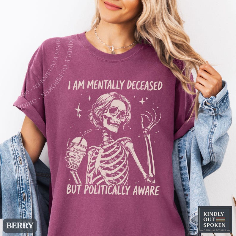 May include: A berry-colored t-shirt featuring a graphic of a skeleton wearing glasses, holding a drink, and surrounded by stars. The text reads I AM MENTALLY DECEASED BUT POLITICALLY AWARE. The shirt is layered under a blue denim jacket.