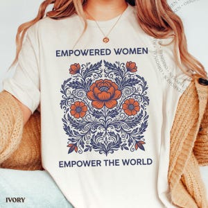 May include: An ivory t-shirt featuring the text EMPOWERED WOMEN EMPOWER THE WORLD in dark blue lettering. The centre graphic displays a detailed floral illustration with orange flowers and dark blue swirling leaves and stems on a light background.