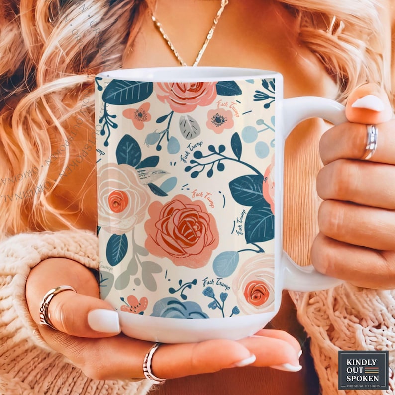 May include: White ceramic mug with a floral pattern in shades of pink, blue, and cream. The design includes roses and leaves, with the phrase "Fuck Trump" repeated throughout. The mug has a white handle.