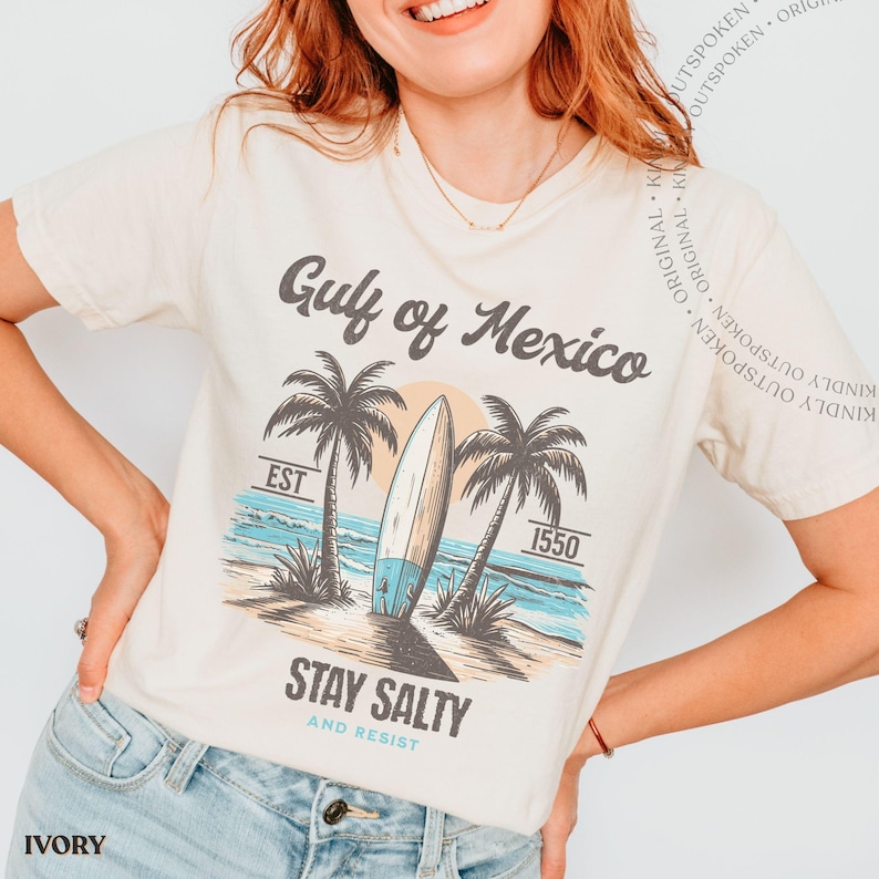May include: An ivory short-sleeve t-shirt featuring a graphic of a surfboard between two palm trees on a beach. The text reads Gulf of Mexico, EST 1550, STAY SALTY AND RESIST. The design uses shades of blue, brown, and tan to depict the coastal scene.