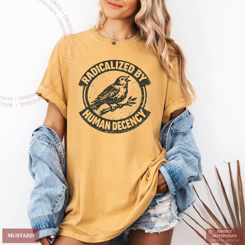 May include: A mustard yellow short-sleeve t-shirt featuring a black circular graphic with a bird perched on a branch in the center. The text RADICALIZED BY HUMAN DECENCY is printed in a bold, distressed font around the circular border.