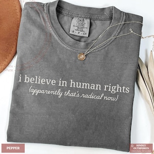 Könnte beinhalten: Ein anthrazitfarbenes Kurzarm-T-Shirt mit Rundhalsausschnitt. Das Shirt trägt den weißen Aufdruck: i believe in human rights (apparently that's radical now). Eine dünne goldfarbene Halskette mit einem kleinen runden Anhänger liegt auf dem Stoff.