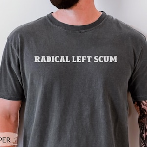 May include: A charcoal grey short-sleeved t-shirt with a crew neckline. The shirt features the text "RADICAL LEFT SCUM" printed in a white, serif font across the center of the chest. The fabric has a washed, vintage-style appearance.
