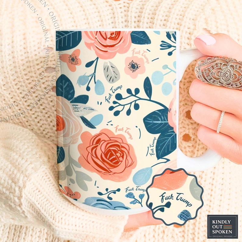 May include: White ceramic mug featuring a floral design in coral, light blue, and grey. The pattern includes roses and the text "Fuck Trump". The mug is held by a person wearing rings.
